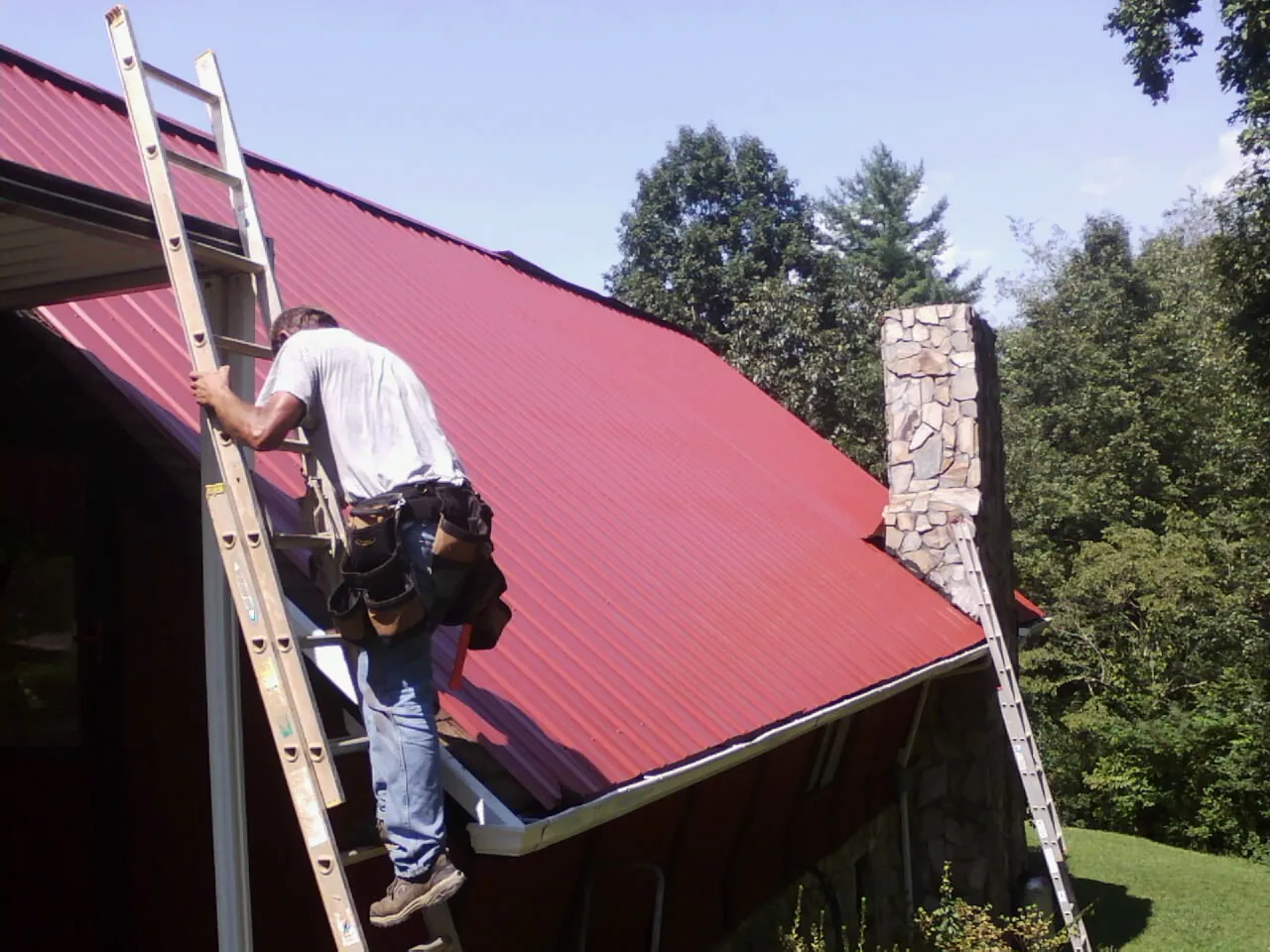 Roofer on a ladder beside a red metal roof and stone chimney — Soffit & Fascia Repair by Roofing Brothers in Burgess